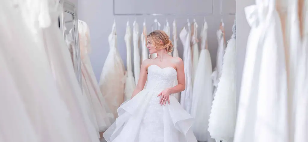 Blonďatá nevěsta si vybírá bílé svatební šaty ve svatebním salonu.